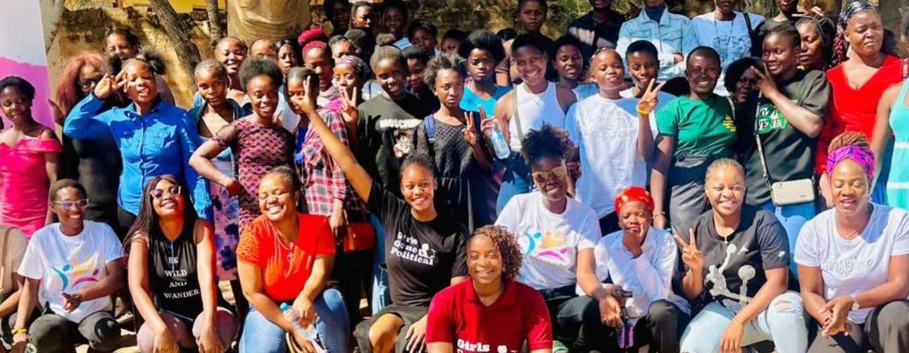 A large group of young women in a rural community in Zambia standing and kneeling outside for a group shot next to the Girls Gone Political banner.