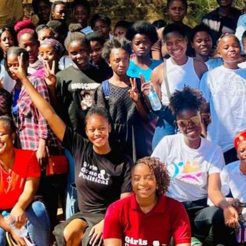 A large group of young women in a rural community in Zambia standing and kneeling outside for a group shot next to the Girls Gone Political banner.