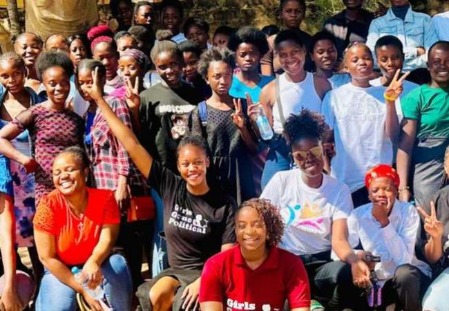 A large group of young women in a rural community in Zambia standing and kneeling outside for a group shot next to the Girls Gone Political banner.