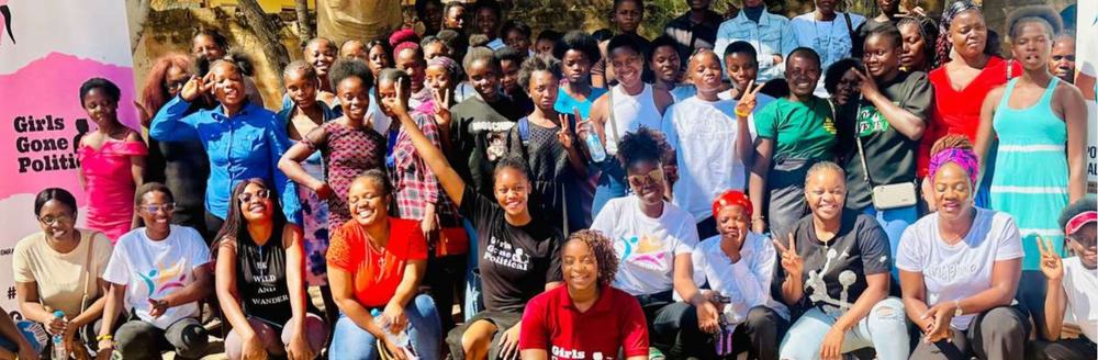 A large group of young women in a rural community in Zambia standing and kneeling outside for a group shot next to the Girls Gone Political banner.