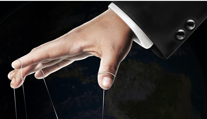 The arm of a men's dark suit with a black background. The hand is holding the control bar of a marionette puppet with strings controlling a plain wooden figure.