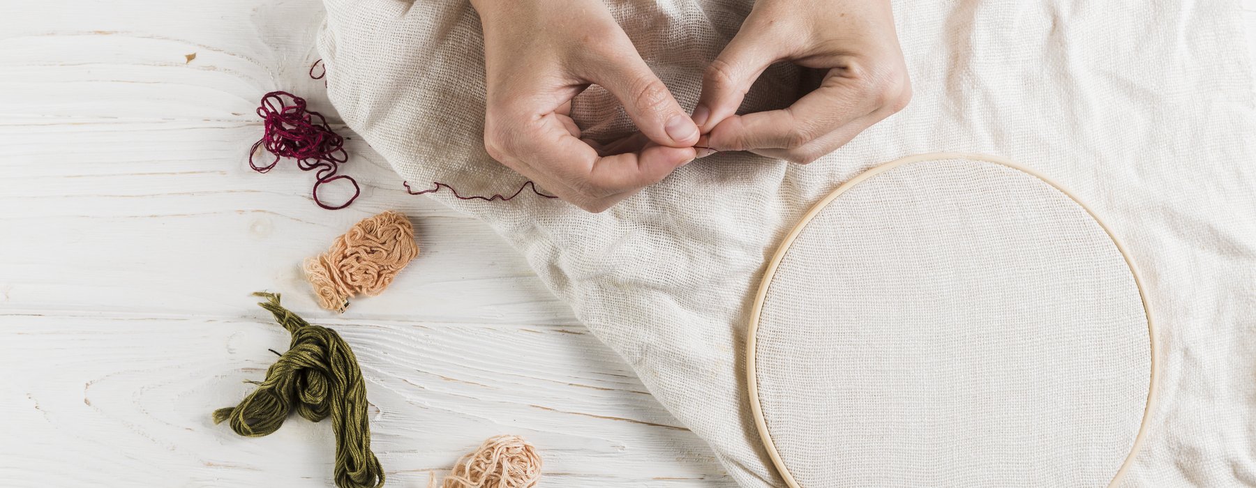 A pair of hands holding a needle and red thread. Behind is a wooden floor that has a white piece of clothing with an embroidery hoop and more threads placed next to it.