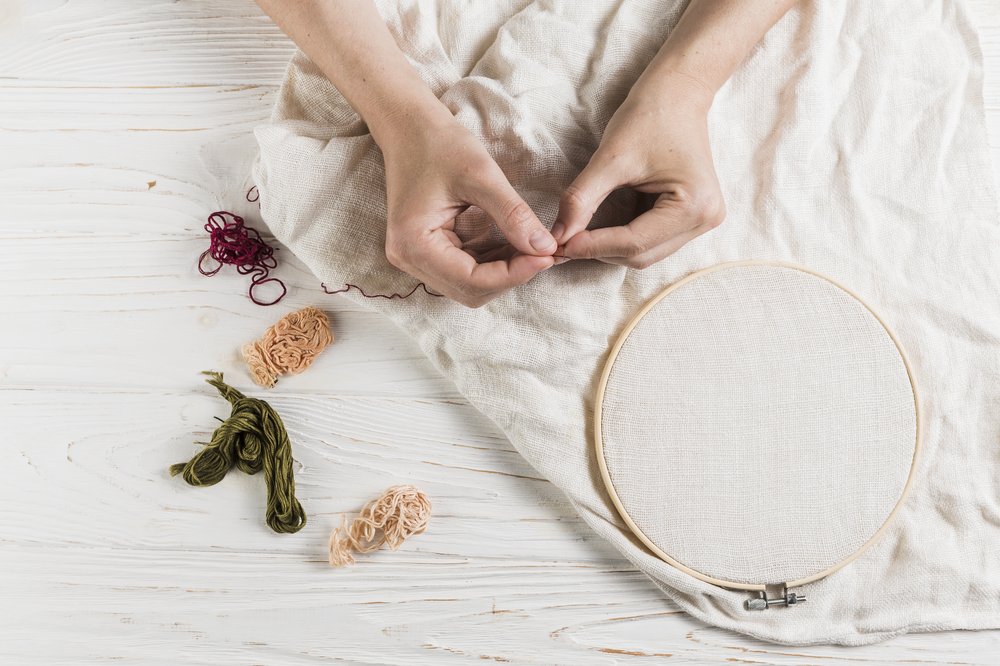 A pair of hands holding a needle and red thread. Behind is a wooden floor that has a white piece of clothing with an embroidery hoop and more threads placed next to it.