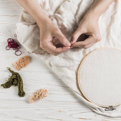 A pair of hands holding a needle and red thread. Behind is a wooden floor that has a white piece of clothing with an embroidery hoop and more threads placed next to it.