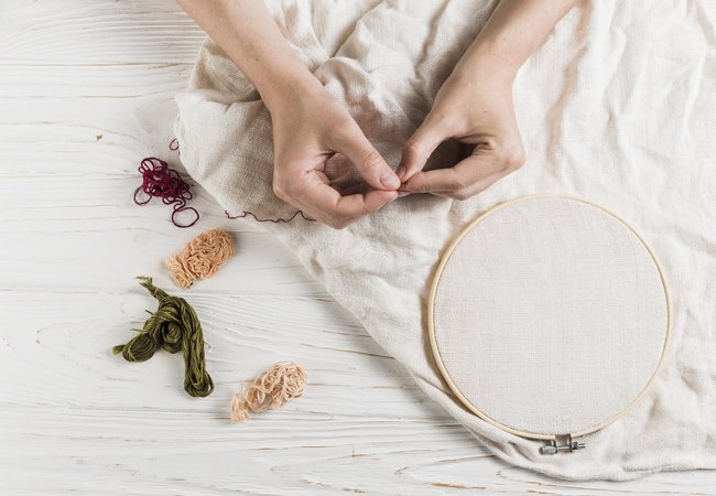 A pair of hands holding a needle and red thread. Behind is a wooden floor that has a white piece of clothing with an embroidery hoop and more threads placed next to it.