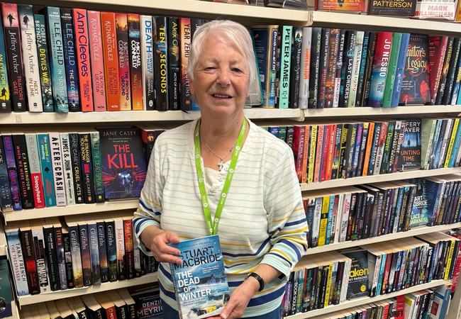 Image of Helen standing in front of a bookshelf holding a book titled 'In the dead of winter'