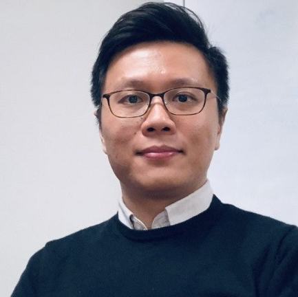 A man with short dark hair and glasses looks into the camera. He is wearing a black jumper with a white shirt.