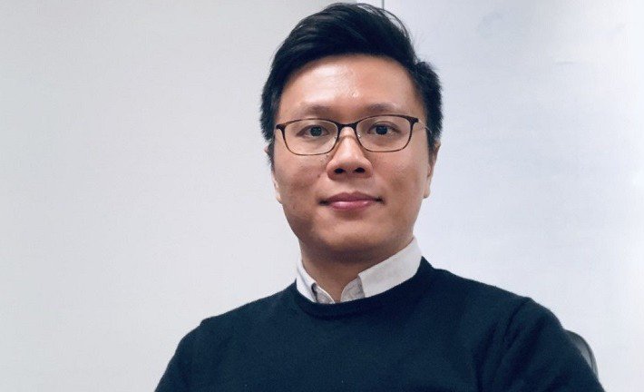 A man with short dark hair and glasses looks into the camera. He is wearing a black jumper with a white shirt.