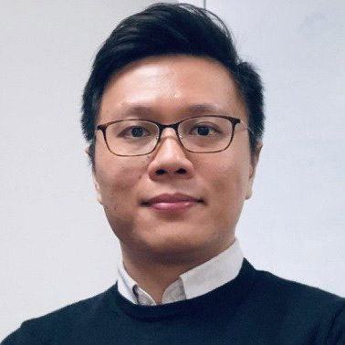 A man with short dark hair and glasses looks into the camera. He is wearing a black jumper with a white shirt.