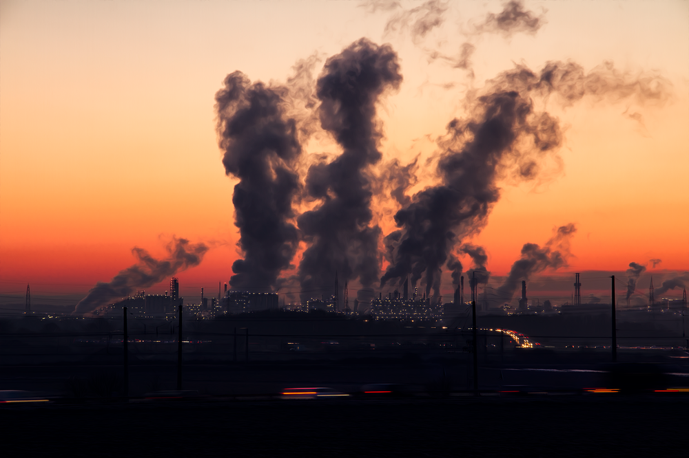 A large factory in silhouette against a clear evening sky with the sunsetting and large plumes of smoke coming out of the factory chimneys.