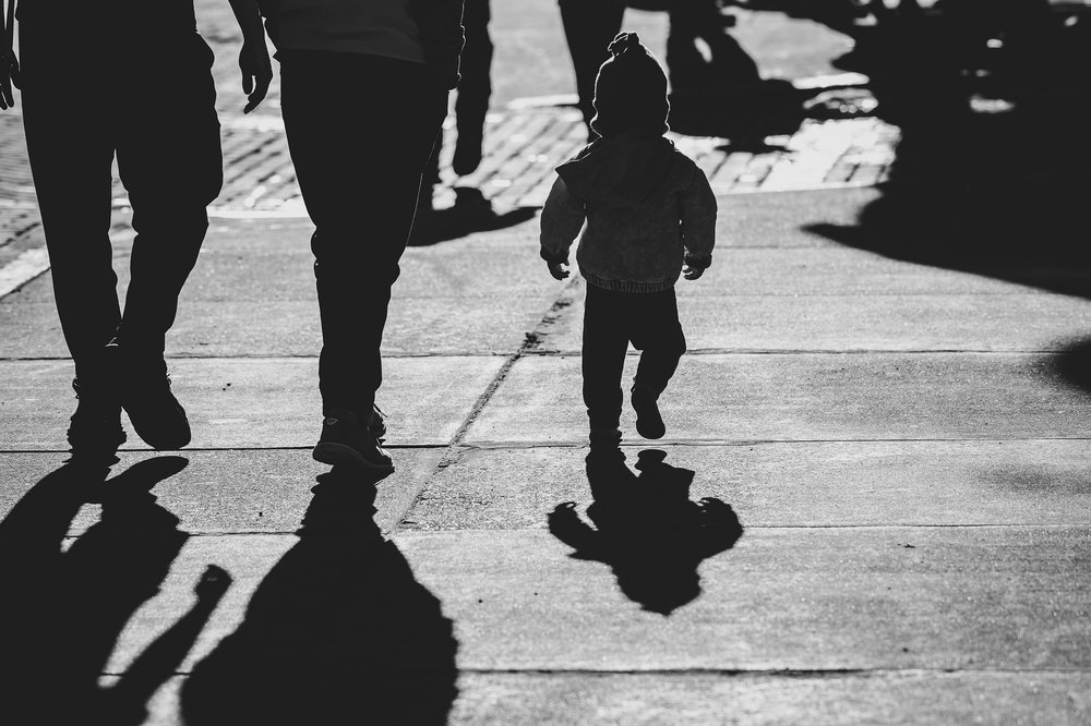A black and white photo of a child walking down the street wearing a winter hat. You can see adults legs walking alongside.