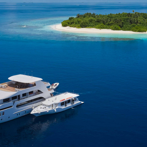 Aerial view of a large luxury yacht and a smaller boat next to it are anchored in a deep blue ocean very near a small island with a sandy beach and green trees