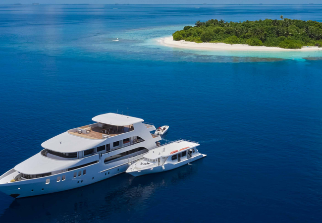 Aerial view of a large luxury yacht and a smaller boat next to it are anchored in a deep blue ocean very near a small island with a sandy beach and green trees