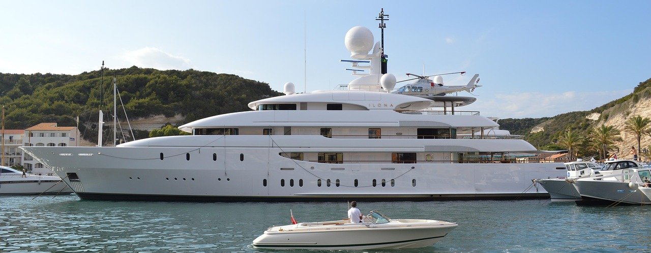 A large white super yacht in a marina with a helicopter on a landing platform near the top of the yacht. In the foreground is a smaller white boat with the man on board looking at the super yacht.