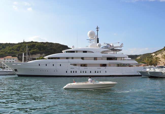 A large white super yacht in a marina with a helicopter on a landing platform near the top of the yacht. In the foreground is a smaller white boat with the man on board looking at the super yacht.