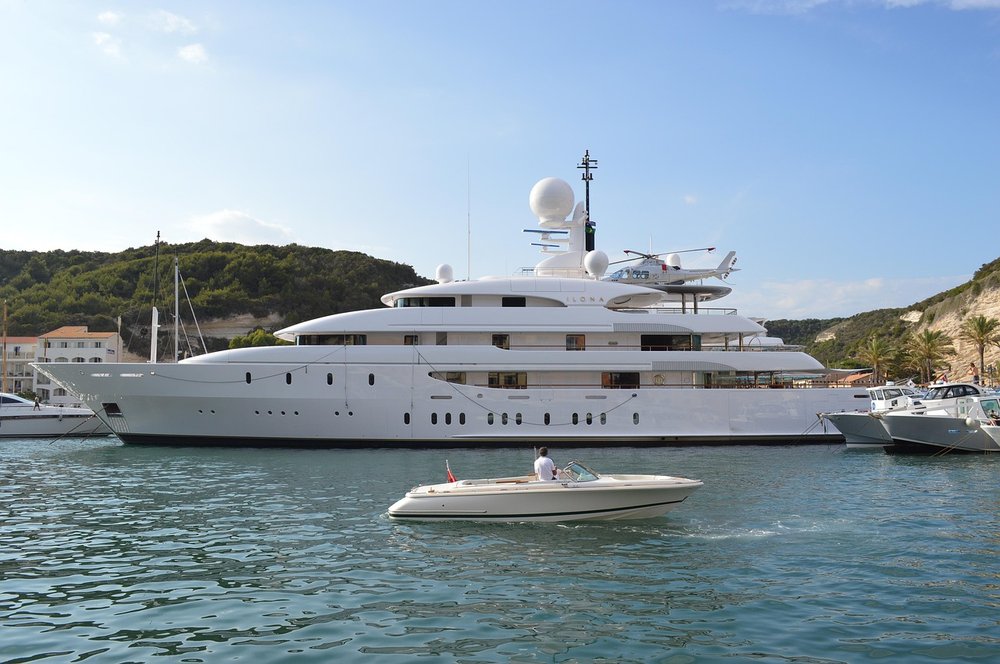 A large white super yacht in a marina with a helicopter on a landing platform near the top of the yacht. In the foreground is a smaller white boat with the man on board looking at the super yacht.