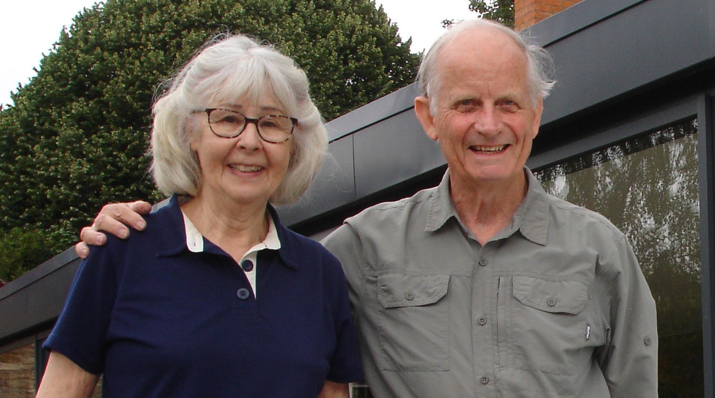 A married elderly couple are standing together outside next to a large window. The husband has his arm around his wife and they are both smiling to the camera.