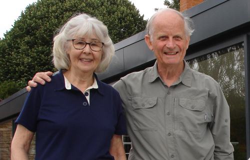 A married elderly couple are standing together outside next to a large window. The husband has his arm around his wife and they are both smiling to the camera.