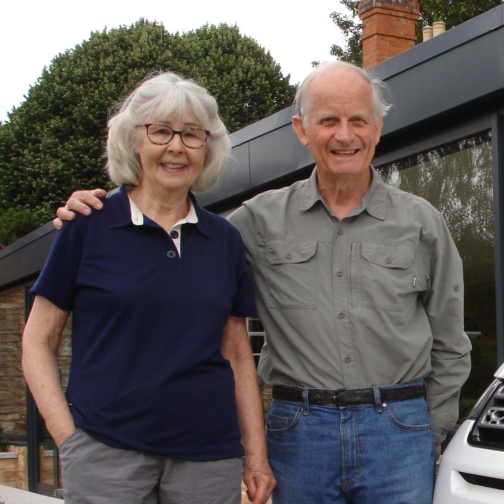 A married elderly couple are standing together outside next to a large window. The husband has his arm around his wife and they are both smiling to the camera.