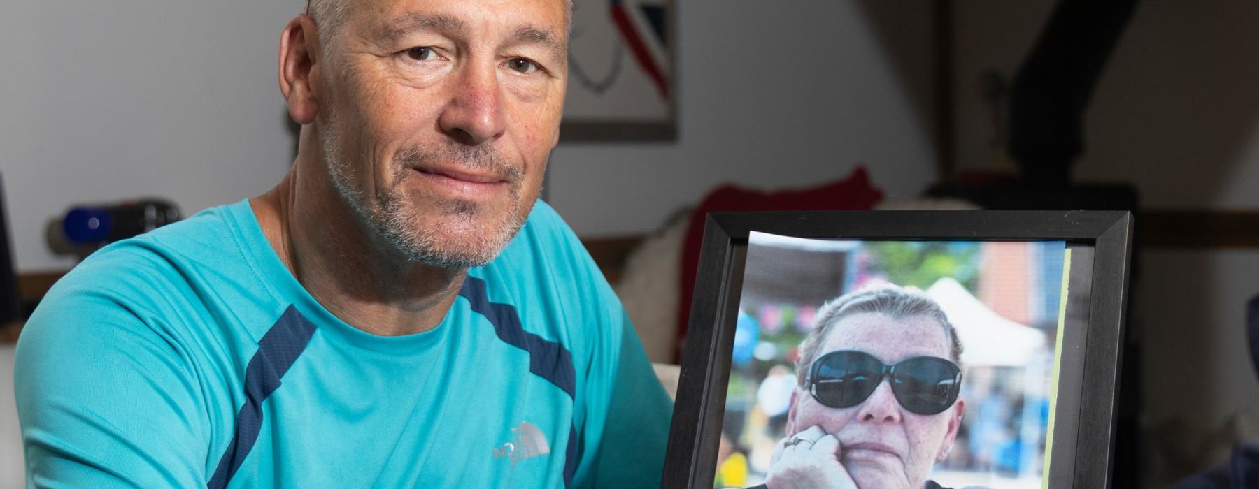 Matt is sat at a dark wood table holding a photograph of his sister Lisa who is wearing sunglasses.