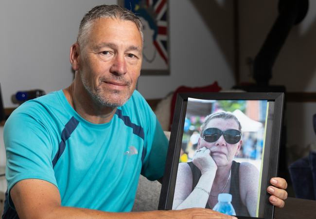 Matt is sat at a dark wood table holding a photograph of his sister Lisa who is wearing sunglasses.