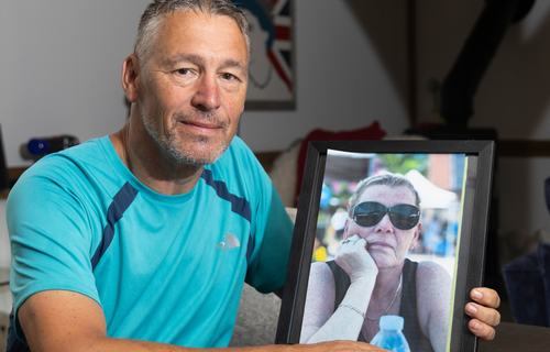 Matt is sat at a dark wood table holding a photograph of his sister Lisa who is wearing sunglasses.