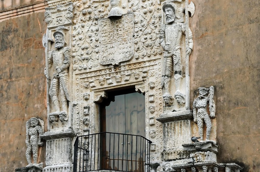 A large stone-carved engraving around a grand wooden door on the outside of a stone building in Mexico with depictions of Spanish military standing on heads with the mouths open like they are screaming