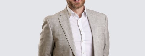 A man with a beard is standing and smiling at the camera. He is wearing a light beige suit jacket over a white dress shirt, with his hands behind his back.