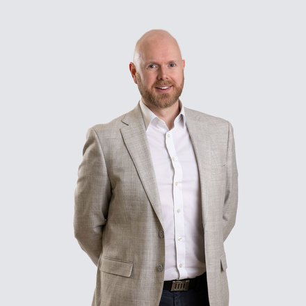 A man with a beard is standing and smiling at the camera. He is wearing a light beige suit jacket over a white dress shirt, with his hands behind his back.