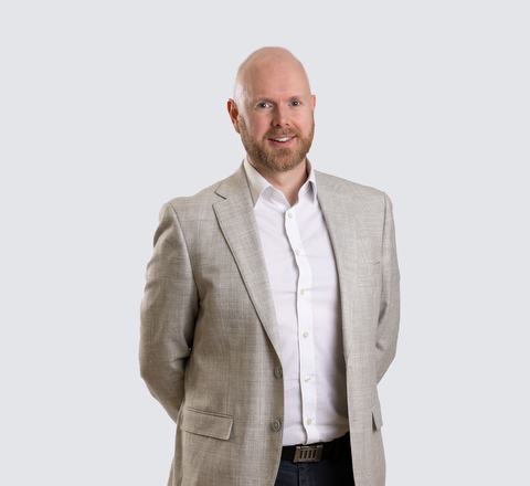 A man with a beard is standing and smiling at the camera. He is wearing a light beige suit jacket over a white dress shirt, with his hands behind his back.
