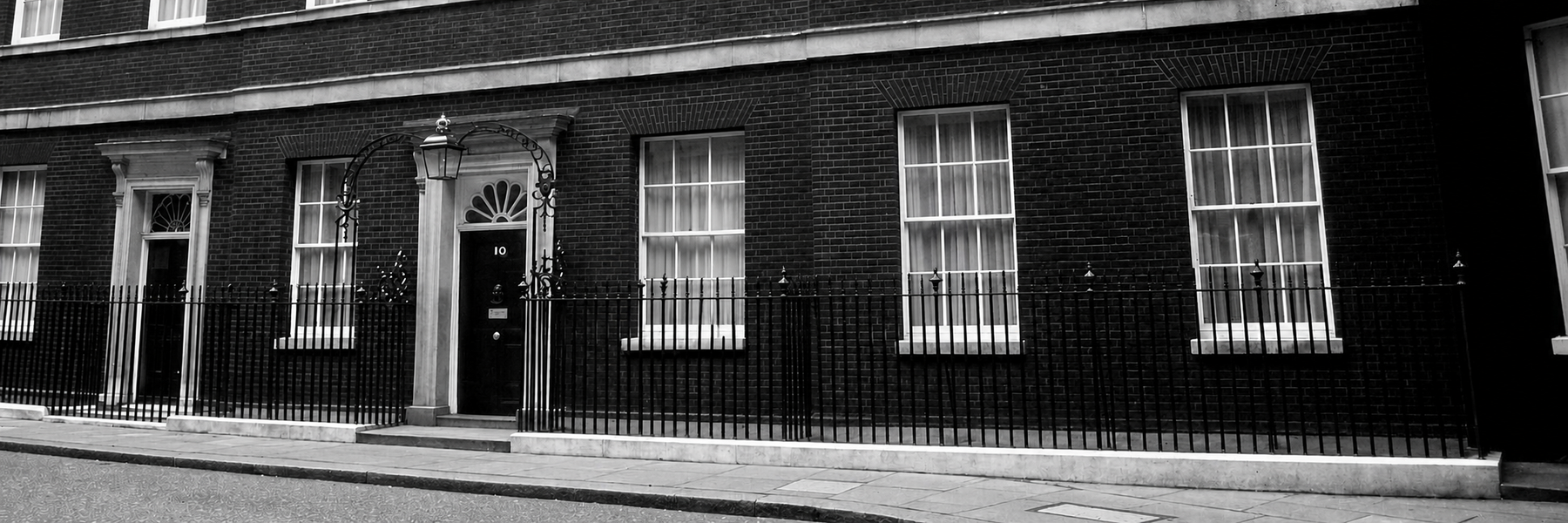 Black-and-white photo of the front of 10 Downing Street, showing the iconic black door, brick façade, sash windows, and iron railings.