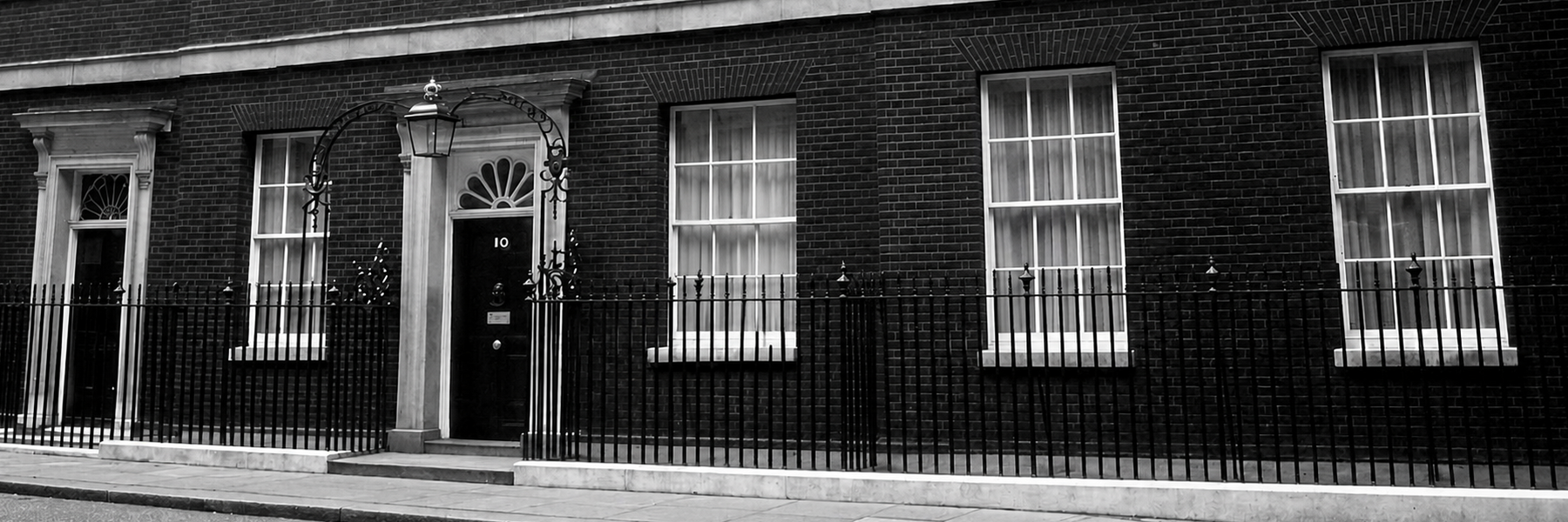 Black-and-white photo of the front of 10 Downing Street, showing the iconic black door, brick façade, sash windows, and iron railings.
