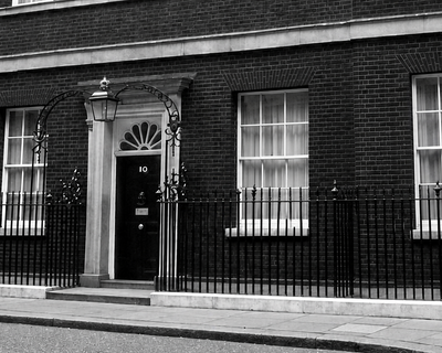 Black-and-white photo of the front of 10 Downing Street, showing the iconic black door, brick façade, sash windows, and iron railings.