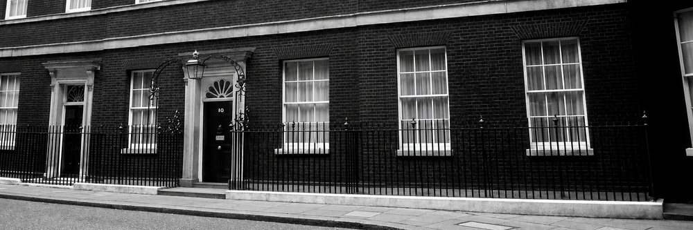 Black-and-white photo of the front of 10 Downing Street, showing the iconic black door, brick façade, sash windows, and iron railings.