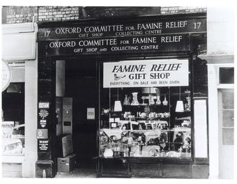 Historic black-and-white photograph of the Oxford Committee for Famine Relief gift shop and collecting centre. The shopfront window is filled with donated goods, with signs reading “Famine Relief Gift Shop” and “Oxford Committee for Famine Relief".