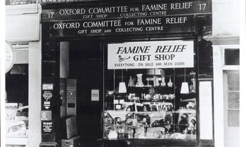 Historic black-and-white photograph of the Oxford Committee for Famine Relief gift shop and collecting centre. The shopfront window is filled with donated goods, with signs reading “Famine Relief Gift Shop” and “Oxford Committee for Famine Relief".