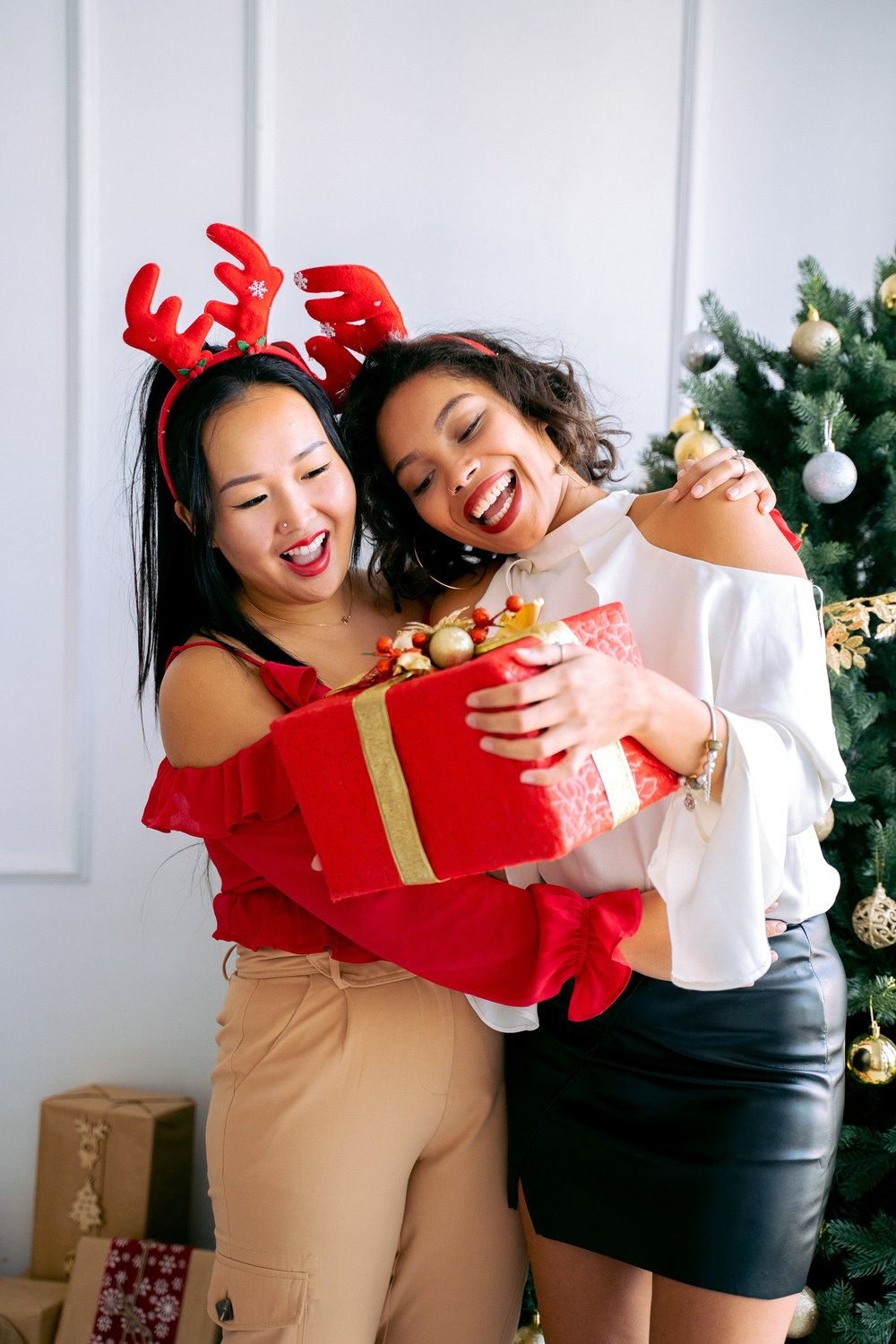 Two female friends hug wearing reindeer hats and holding a big red wrapped Christmas present with a big bow