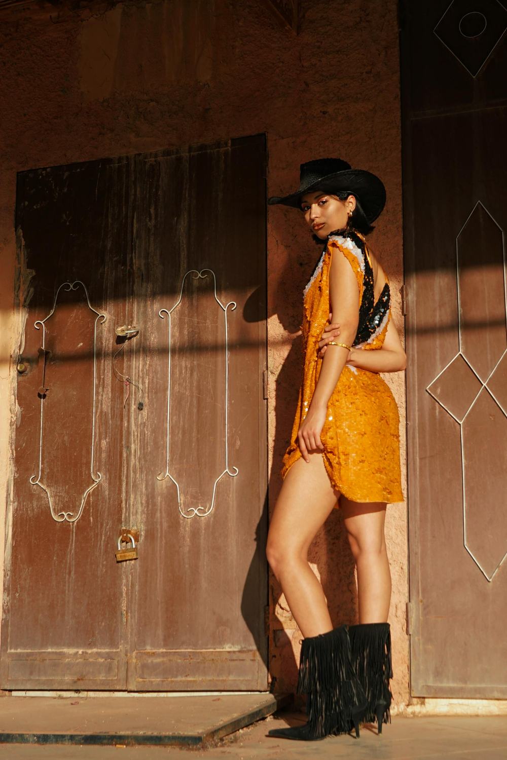 A person wearing an orange mini dress with a black cowboy hat and black fringed cowboy boots.