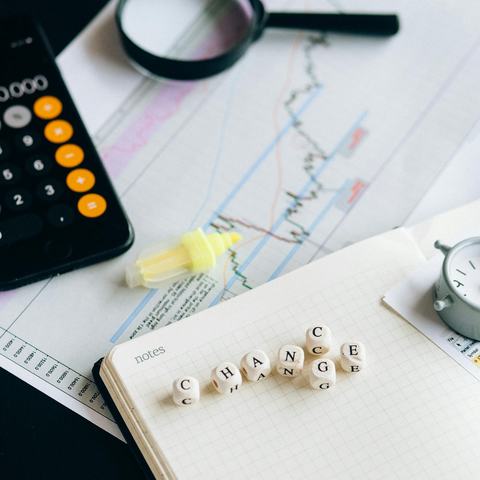 A desk with a sheet of paper showing a line graph with lots of peaks and troughs, a magnifying glass and coloured highlighters. There is also a pad with scrabble letters spelling C-H-A-N-G-E and a single letter C just above the G.