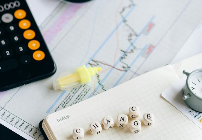 A desk with a sheet of paper showing a line graph with lots of peaks and troughs, a magnifying glass and coloured highlighters. There is also a pad with scrabble letters spelling C-H-A-N-G-E and a single letter C just above the G.
