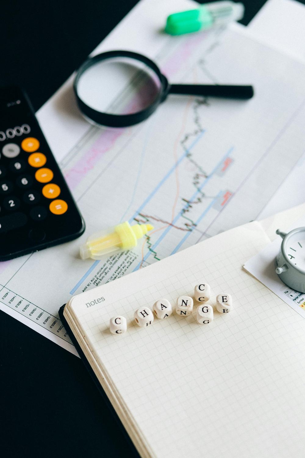 A desk with a sheet of paper showing a line graph with lots of peaks and troughs, a magnifying glass and coloured highlighters. There is also a pad with scrabble letters spelling C-H-A-N-G-E and a single letter C just above the G.