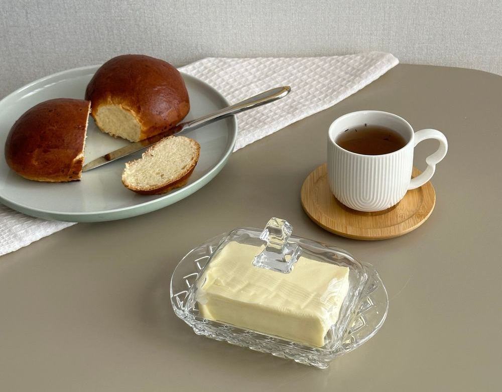 Butter in a clear glass butter dish on a table pictured next to a bread roll and a cup of coffee.