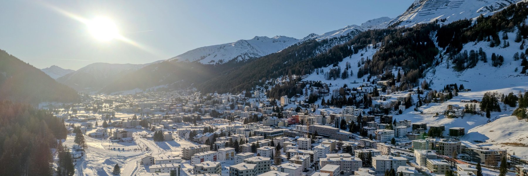 An aerial view of a winter city surrounded by snowy mountains, with bright sunlight casting long shadows across buildings and slopes.