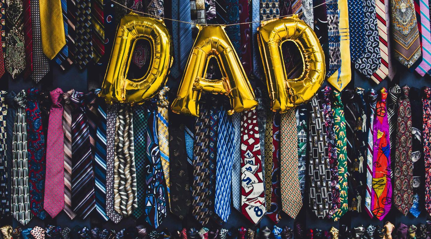 Gold balloons spelling out DAD, on a background of funky pattern ties all hung up. Two rows of around 40 ties.