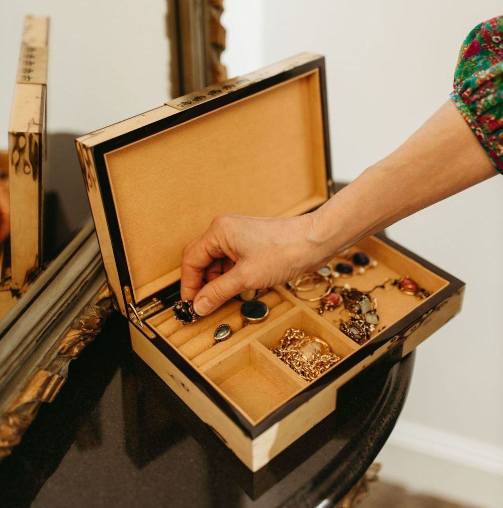 An open wooden jewellery box on an antique black and gold table with a hand reaching for the jewellery inside the box.