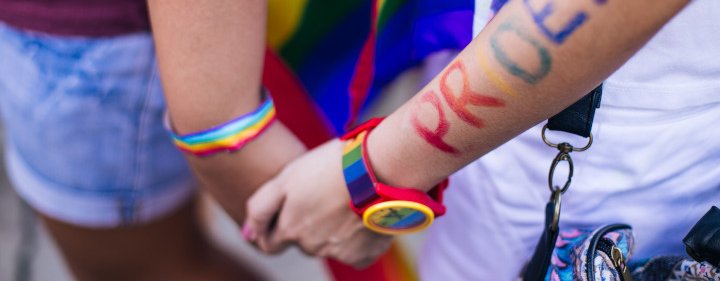 Two people holding hands. A rainbow ribbon is wrapped around one person's wrist and on the other person's wrist is a rainbow watch with the word PRIDE painted in rainbow colours on the arm