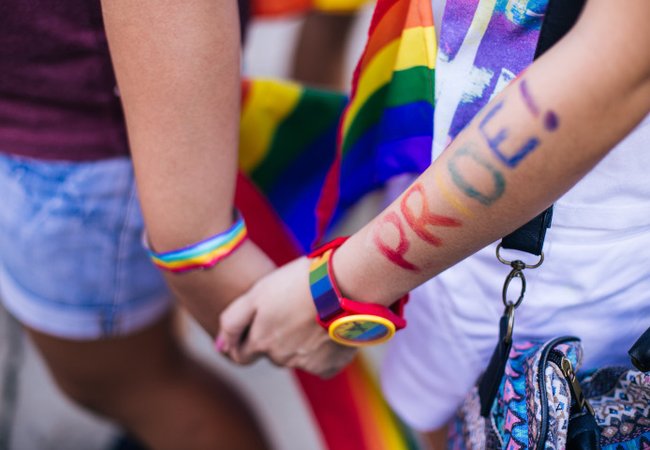 Two people holding hands. A rainbow ribbon is wrapped around one person's wrist and on the other person's wrist is a rainbow watch with the word PRIDE painted in rainbow colours on the arm