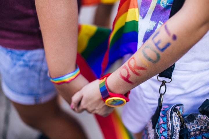 Two people holding hands. A rainbow ribbon is wrapped around one person's wrist and on the other person's wrist is a rainbow watch with the word PRIDE painted in rainbow colours on the arm