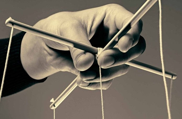 A black and white photo of a hand holding the control bar of a marionette puppet with strings. The puppet is not shown in the picture.