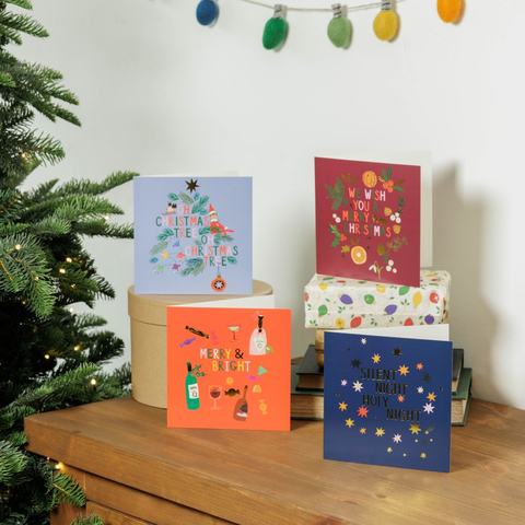Four festive Christmas cards beside a decorated tree, featuring seasonal greetings, gifts and holiday décor.